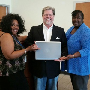 smiling man holding a laptop flanked by two smiling women